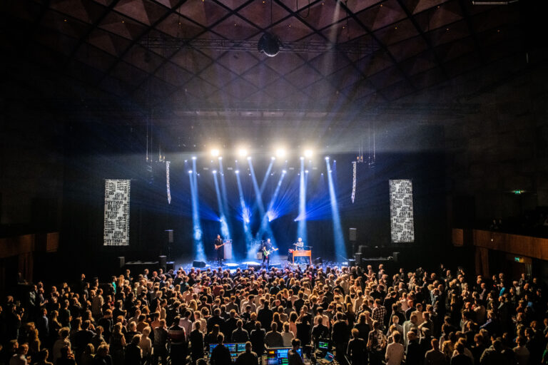 Een liveconcert met een groot publiek, verlicht door spots en podiumverlichting.