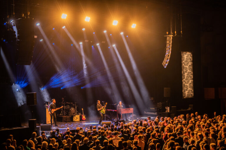 Een liveband optredend op een podium met een menigte en kleurrijke verlichting.