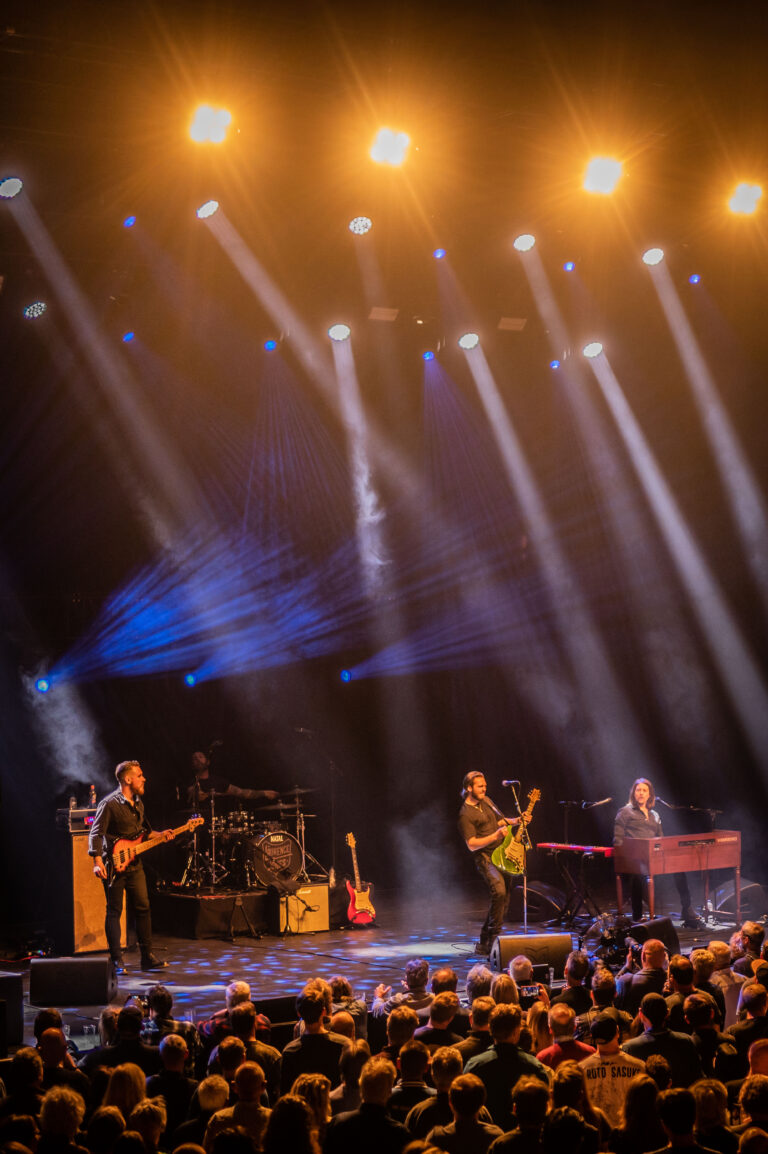 Een live band speelt op het podium voor een enthousiast publiek, met opvallende lichten die de scène verlichten.