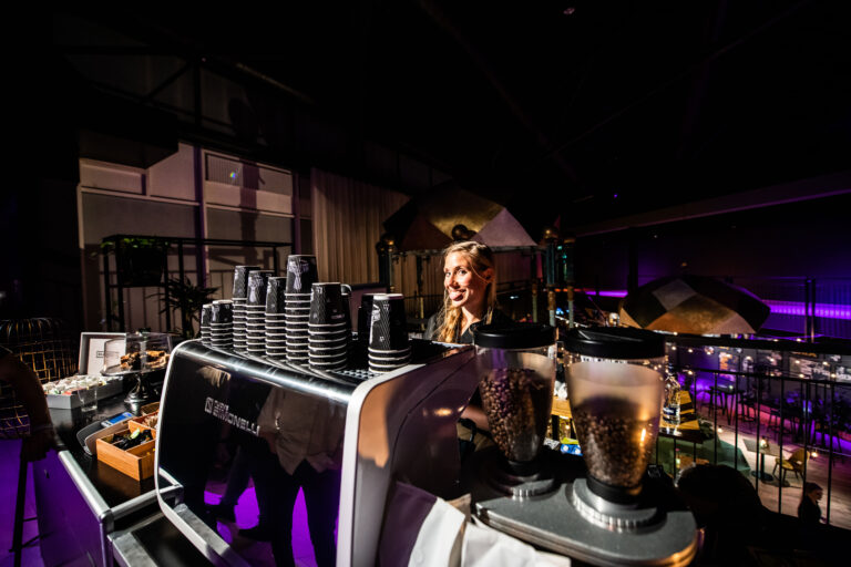 Een barista staat achter een koffiebar met gestapelde bekers en koffieapparatuur in een sfeervolle omgeving.