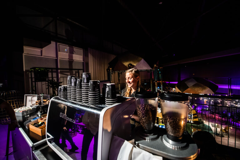 Een barista staat achter een koffiezetmachine, omgeven door stapels bekers in een sfeervolle, verlichte ruimte.