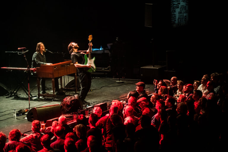Een muzikant speelt gitaar op het podium, terwijl een collega achter een keyboard zit en een enthousiast publiek toekijkt.