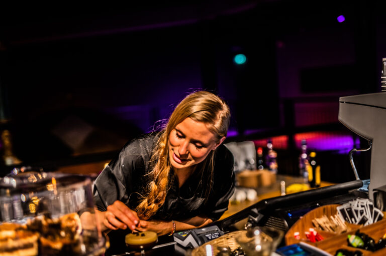 Een vrouw met een lange vlecht werkt aan het bereiden van drankjes achter een bar in een sfeervolle setting.