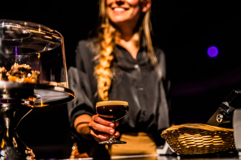 Een vrouwelijke serveerster houdt een glas met een donker drankje vast, terwijl ze naast een glazen taartplateau staat.