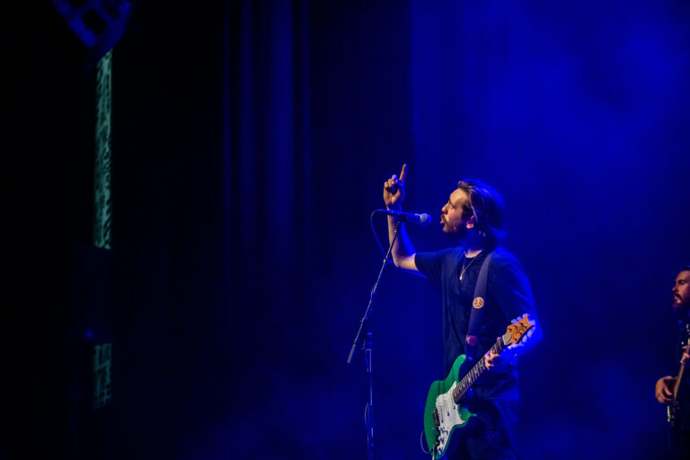 Een muzikant met een groene gitaar staat op een podium, terwijl hij zingt in een blauw verlichte omgeving.