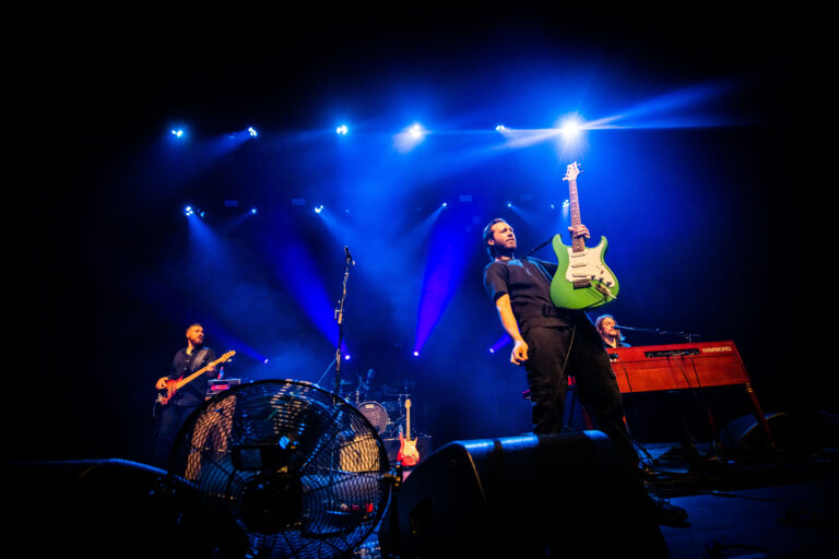 Een band speelt live op het podium, met een muzikant die een groene gitaar omhoog houdt onder blauwige lichten.