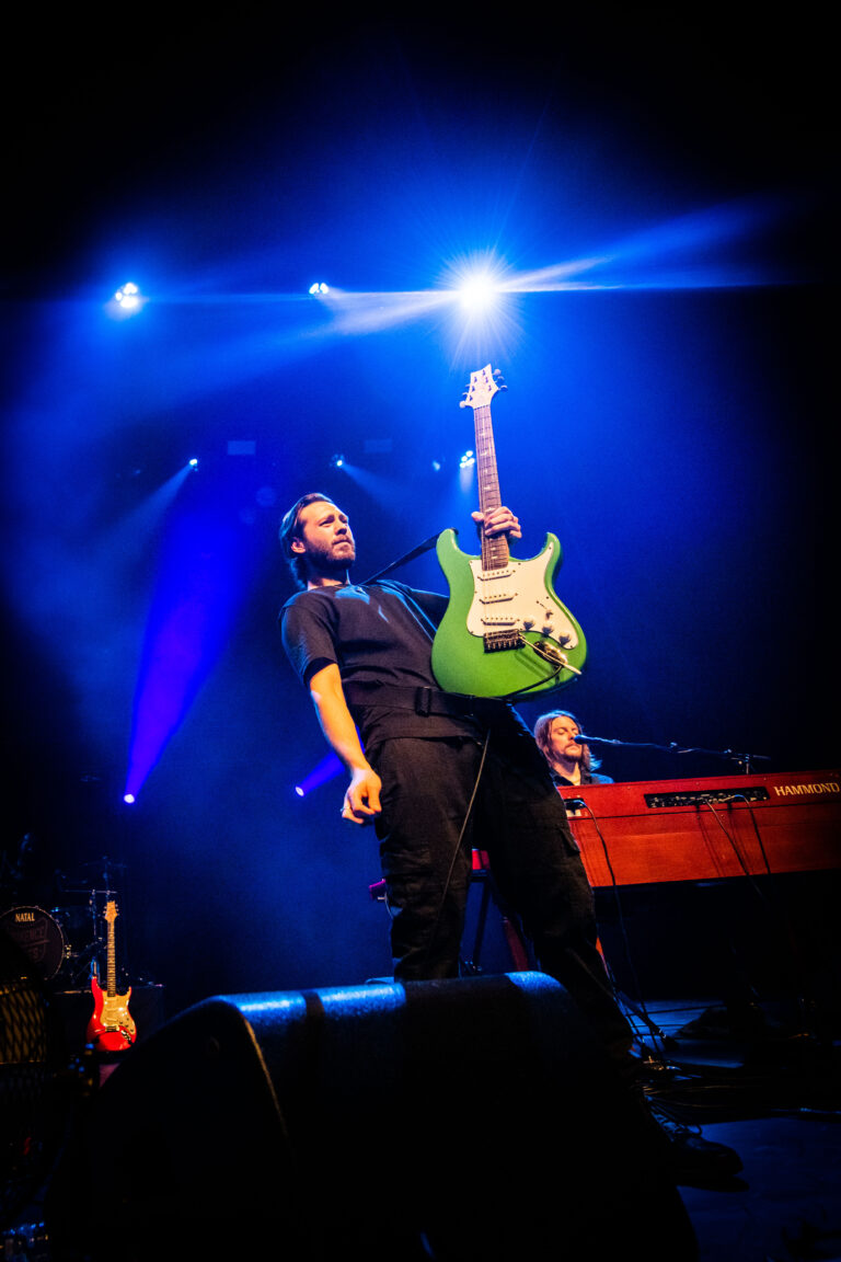 Een muzikant op het podium speelt een groene gitaar onder felle blauwe lichten.