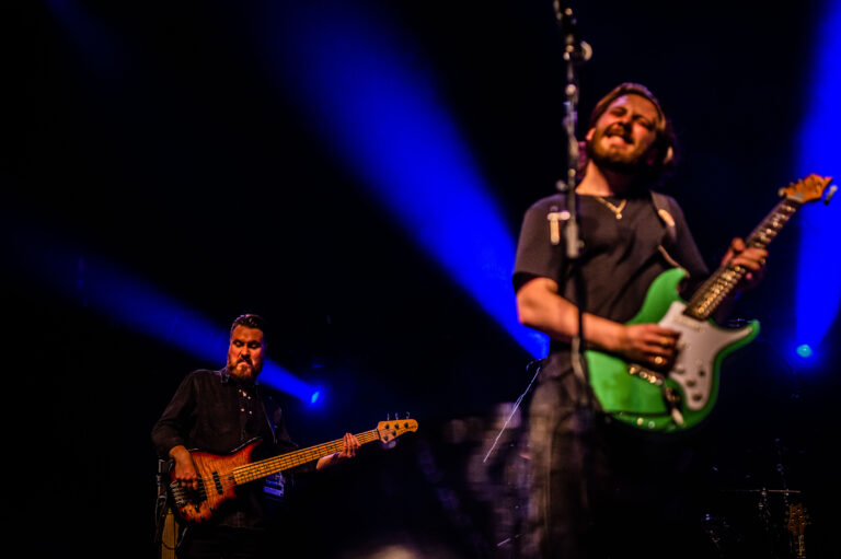 Twee muzikanten op een podium, een met een groene gitaar en de ander met een basgitaar, onderlichte met blauwe lichten.