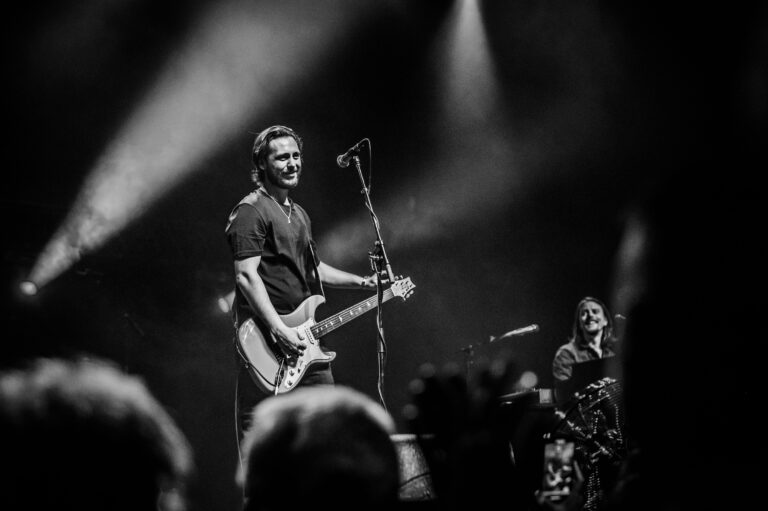 Een muzikant speelt gitaar op het podium terwijl hij naar het publiek kijkt, omringd door stage-licht.