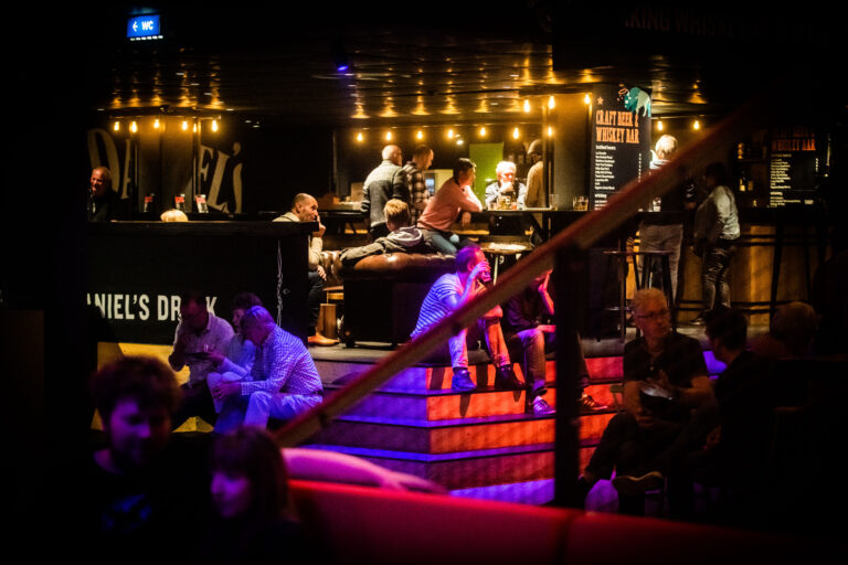 Een sfeervol interieur van een bar met bezoekers die ontspannen op banken zitten en enkele mensen die aan de bar staan.