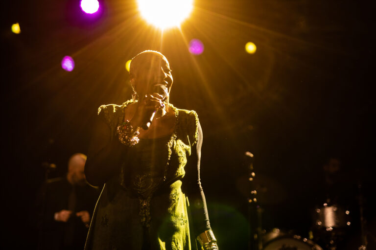 Een zangeres met een microfoon, verlicht door een krachtige spotlicht, staat op een podium tijdens een optreden.