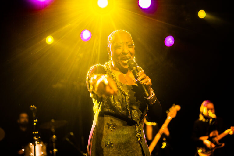 Een zangeres staat op het podium en zingt met een microfoon in de hand, terwijl kleurrijke lichten om haar heen schijnen.