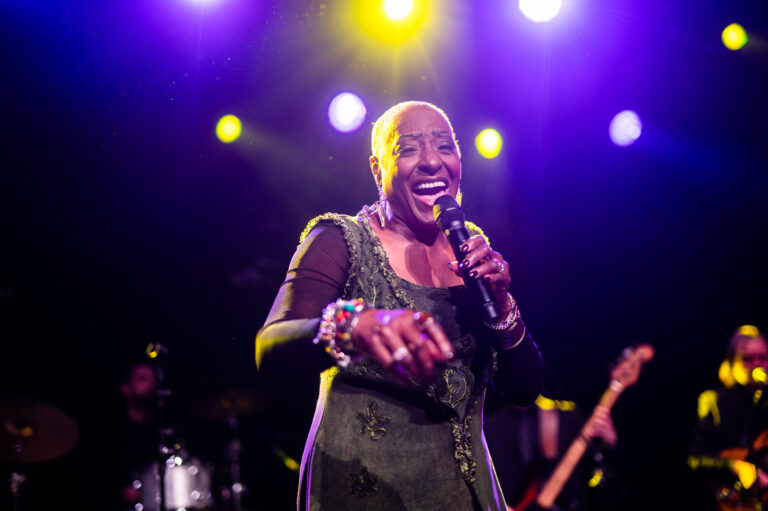 Een vrouwelijke zangeres met een korte shaved head, gekleed in een groene outfit, staat op het podium en zingt met een microfoon in de hand.