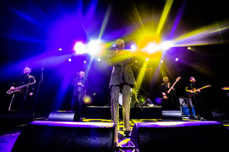 Een muzikale band staat op een podium met felgekleurde lichten tijdens een optreden.