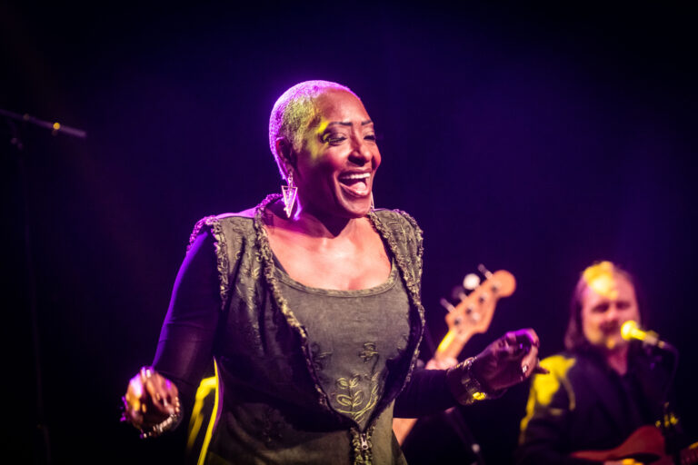 Een zangeres met een kort geschoren hoofd en een vrolijke uitstraling, performing op een podium met muzikanten op de achtergrond.