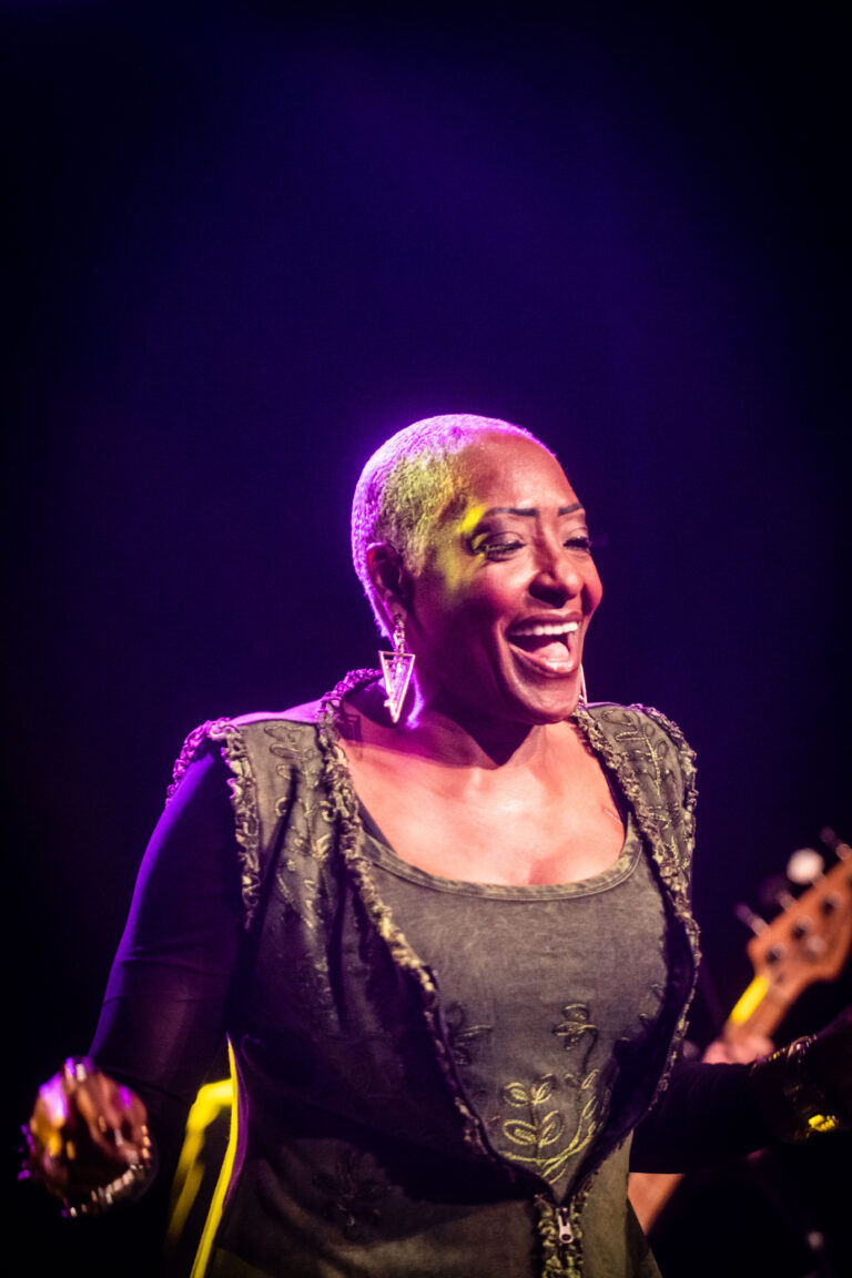 Een zangeres met een vrolijke uitstraling, gekleed in een zwart topje, staat op het podium met een kleurrijke verlichting achter haar.