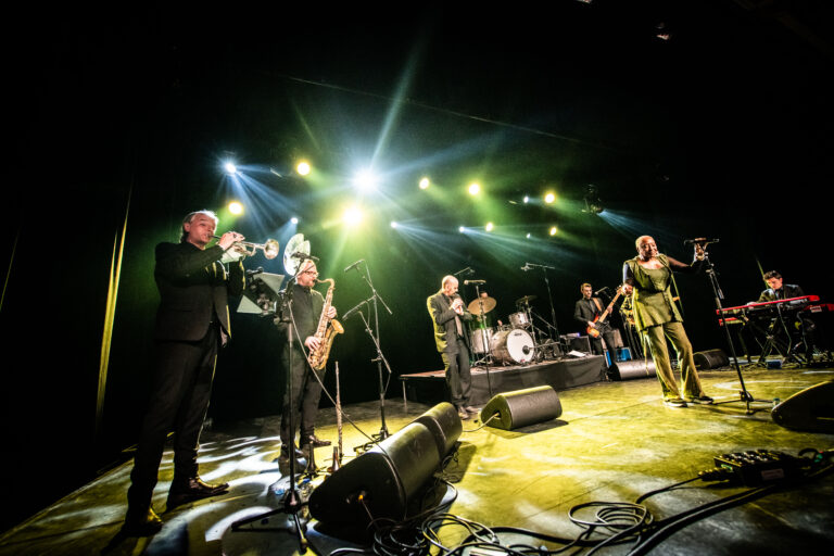 Een band met zes muzikanten voert een live optreden uit op een podium, met kleurrijke verlichting en verschillende instrumenten.