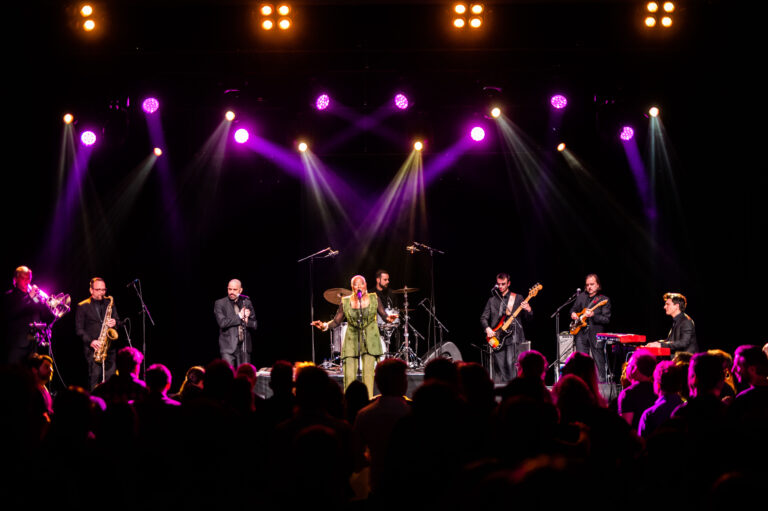 Een band performt live op een podium met kleurrijke verlichting, terwijl het publiek kijkt.