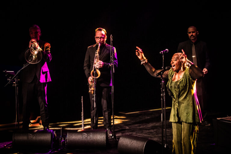 Een zangeres met een groene outfit staat op een podium, omringd door vier muzikanten met blaasinstrumenten, terwijl ze een optreden geven.