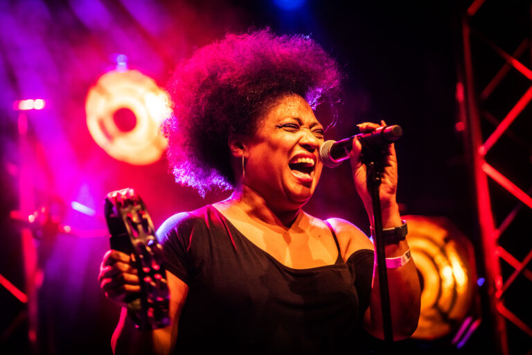 Een muzikante met een afro-haarstijl zingt krachtig in een live optreden, terwijl ze een tamboerijn vasthoudt, omringd door kleurrijke lichten.