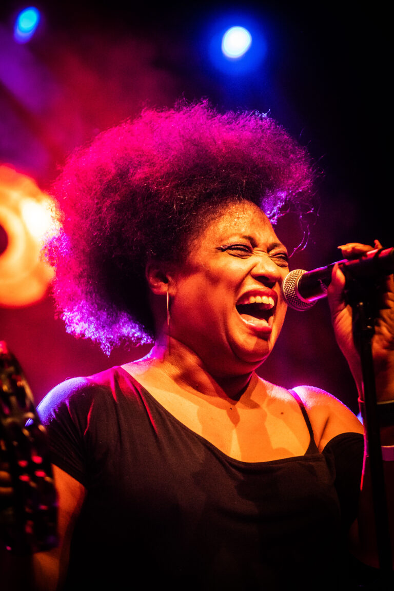 Een vrouw met een volle afro zingt in een microfoon terwijl ze wordt verlicht door kleurrijke lichten op een podium.