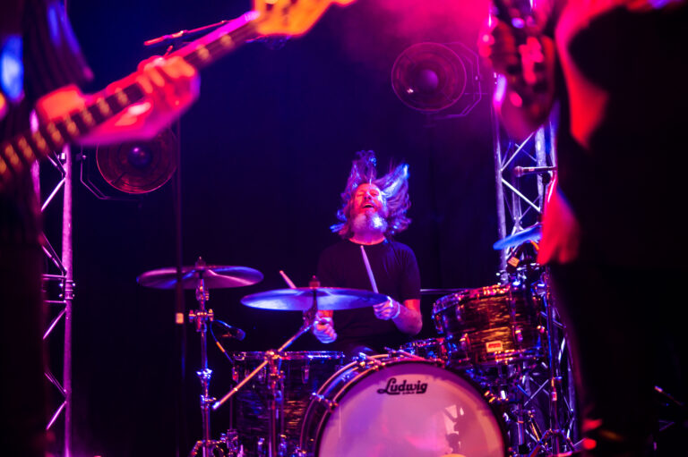 Een drummer speelt op een drumstel terwijl kleurrijke lichten de scène verlichten.