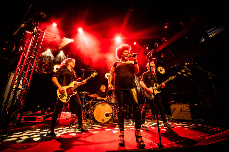 Een band staat op het podium, met felrode lichten en een energieke zangeres in het middelpunt.