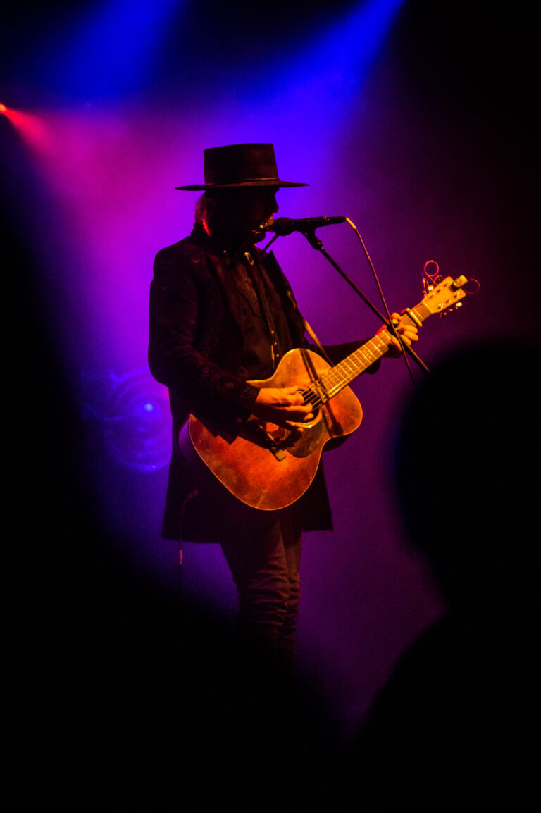 Een muzikant in een hoed speelt gitaar op een podium met kleurrijke verlichting.