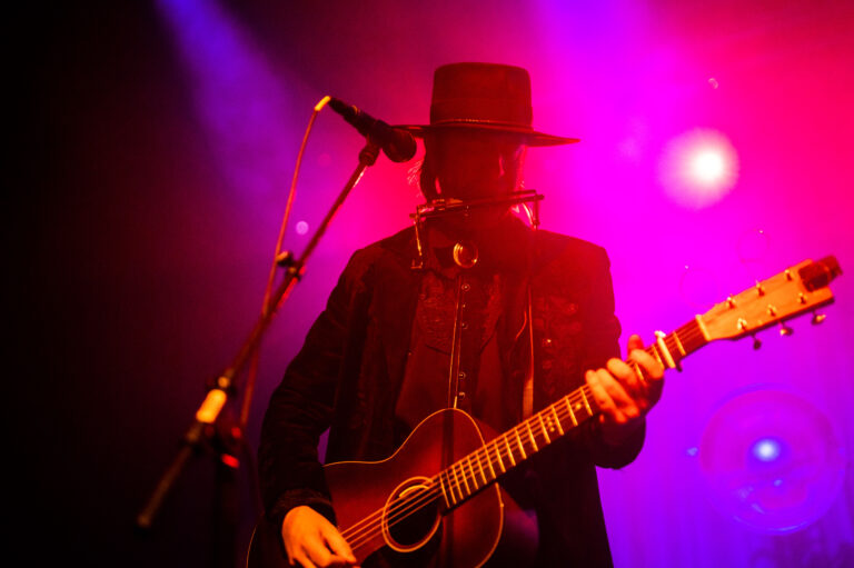 Een muzikant in een zwart kostuum en hoed speelt gitaar op een podium met paarse en roze verlichting.