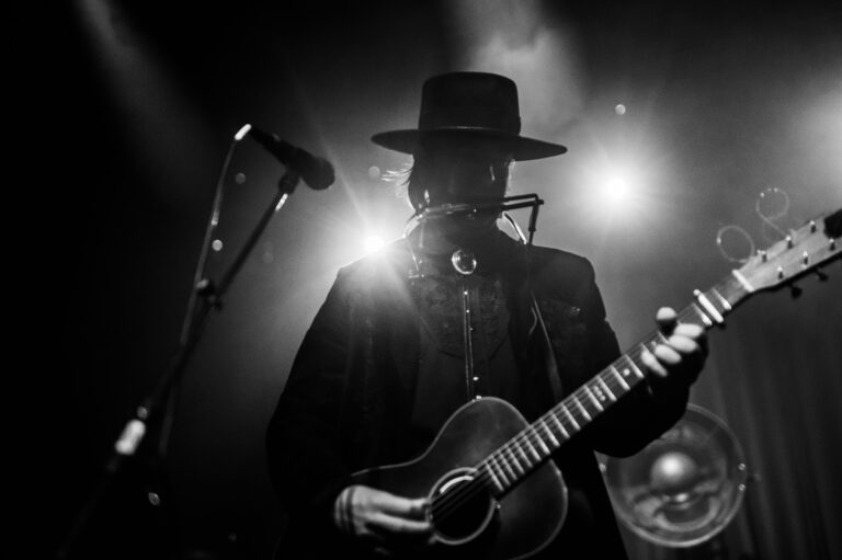 Een silhouet van een muzikant in een hoed die een gitaar bespeelt, omringd door sfeervolle lichten op het podium.
