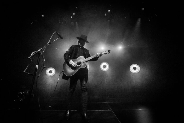 Een silhouet van een muzikant met een gitaar, terwijl hij optreedt op een podium met fel licht.
