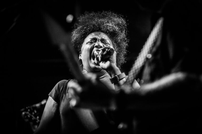 Een zangeres met een volle bos haar zingt intensief in een zwart-wit concertsetting.