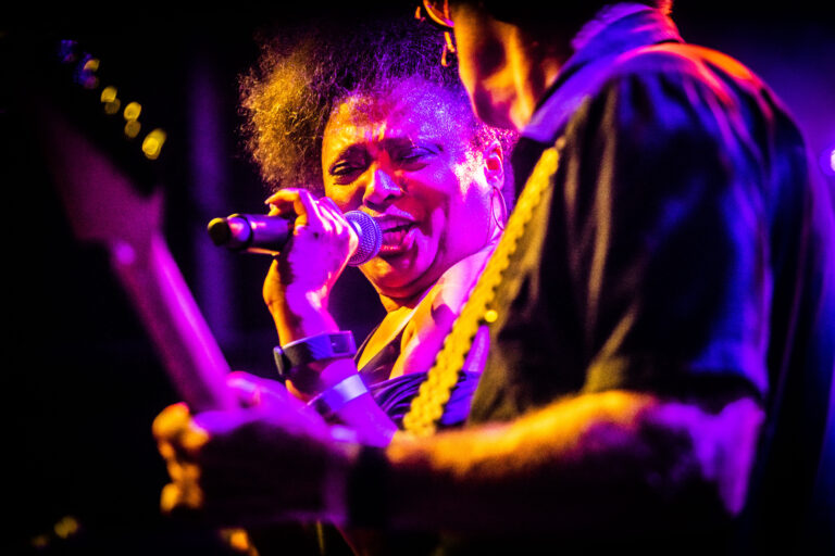 Een zangeres met Afro-haar performt op een podium, terwijl een muzikant in de achtergrond een gitaar vasthoudt.