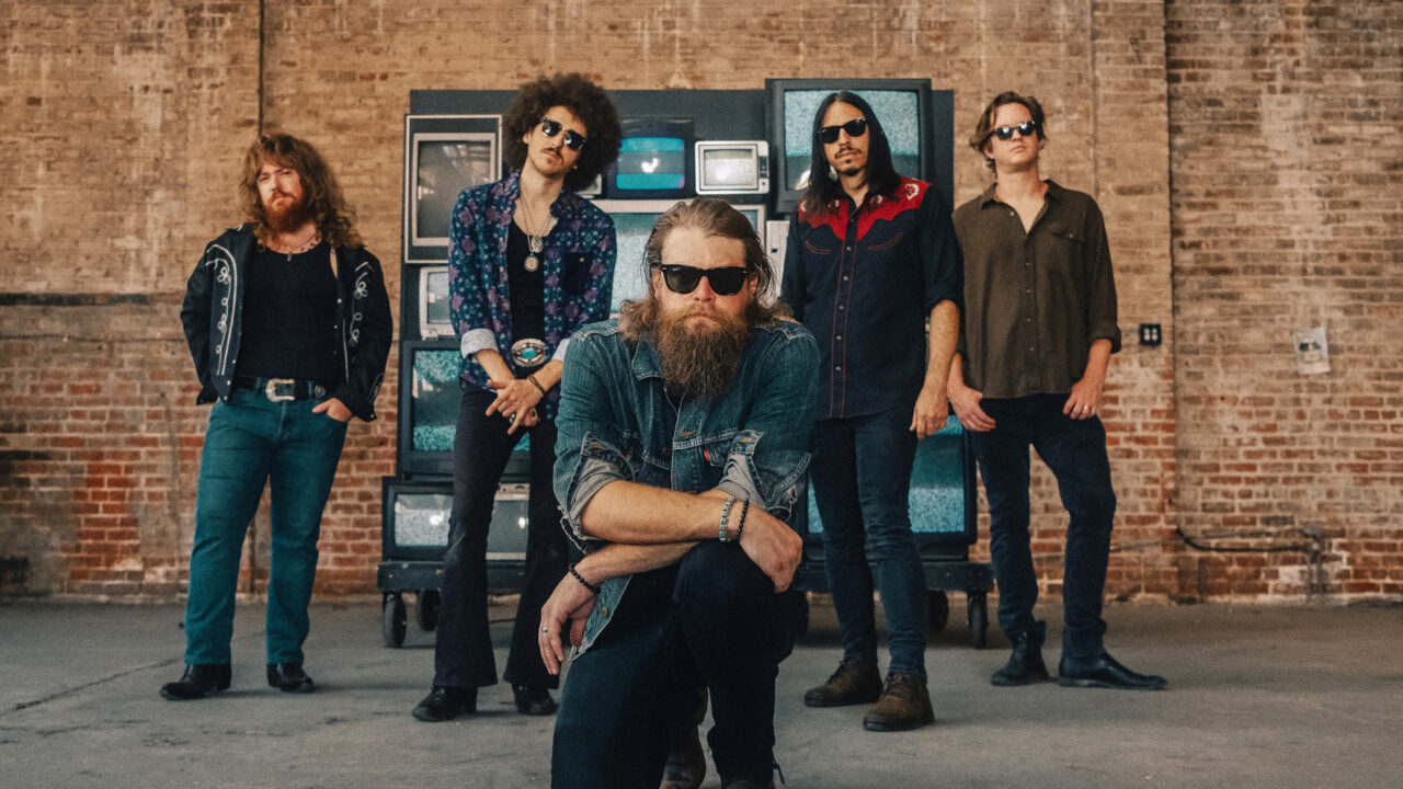 De Amerikaanse bluesrockband Robert Jon & The Wreck poseert in een industriële ruimte, met de zanger vooraan knielend en de andere bandleden erachter, allemaal met zonnebrillen en rock-'n-roll kleding.