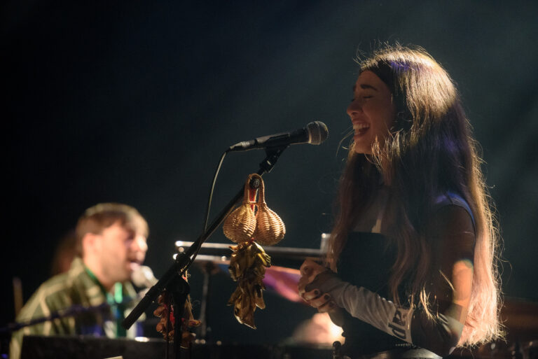 Een vrouwelijke muzikant zingt met enthousiasme achter een microfoon, terwijl een mannelijke muzikant op de achtergrond speelt.