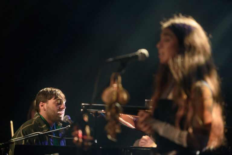 Twee muzikanten voeren een live optreden uit, waarbij één zingt en de ander op een piano speelt, omringd door zachte verlichting.