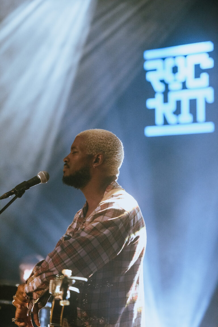 Een muzikant speelt op een podium met een microfoon en bijzondere lichteffecten op de achtergrond.