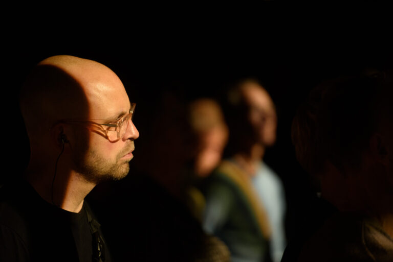 Een man met een kaal hoofd en een bril staat in aanmerking met donkere schaduw op de achtergrond, terwijl andere figuren vaag zichtbaar zijn.