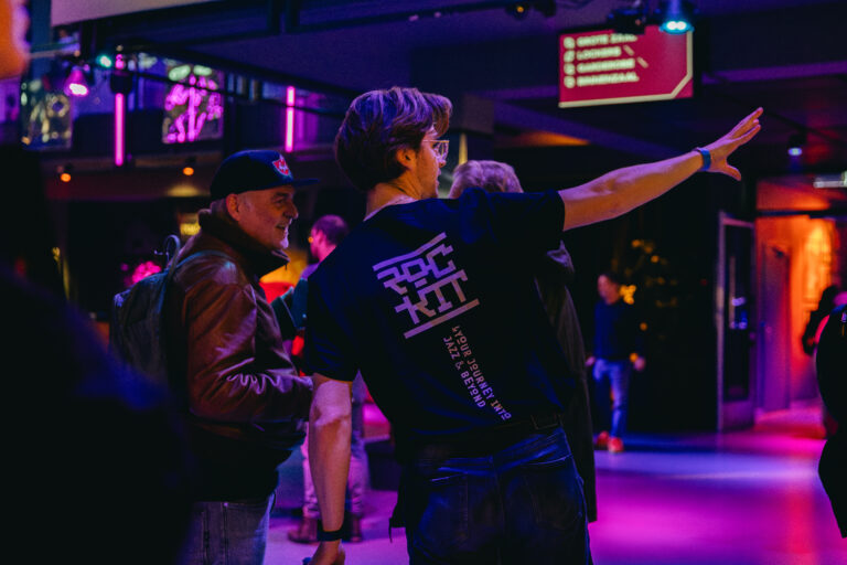 Een jongeman in een zwart t-shirt wijst naar iets terwijl hij zich bevindt in een levendig verlichte ruimte met mensen om hem heen.