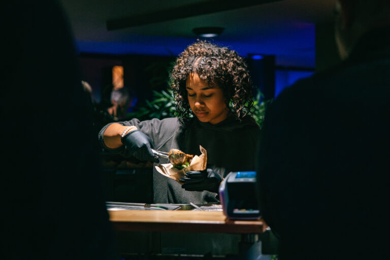 Een persoon met krullend haar, gekleed in een donkere hoodie en handschoenen, serveert voedsel aan een tafel onder gedimd licht.