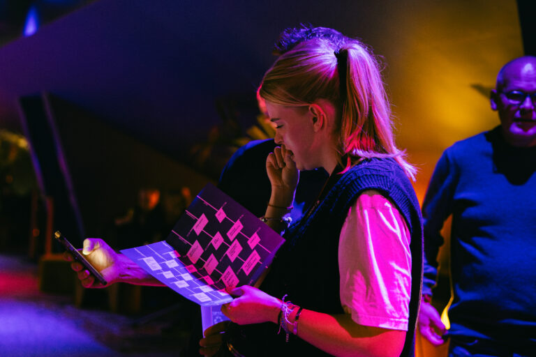 Een persoon met een paardenstaart bekijkt een plattegrond terwijl ze in een kleurrijke, sfeervolle omgeving staat.