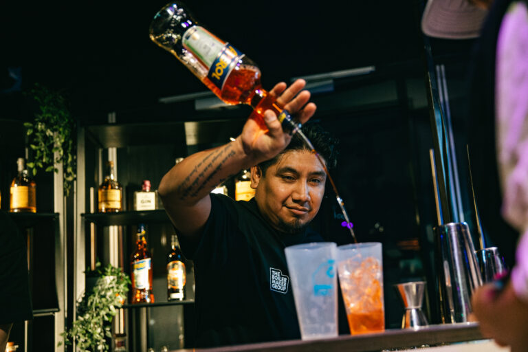 Een barman giet een drankje in een glas achter de bar, omringd door flessen alcohol.