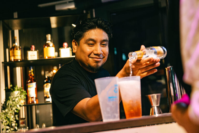 Een barman schenkt een drankje in een glas achter de bar, omringd door flessen sterke drank.