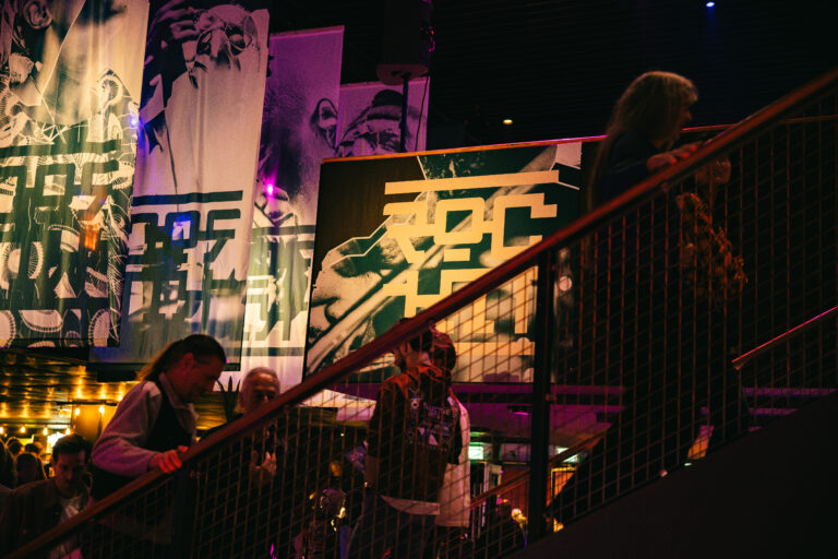 Bezoekers lopen een trap af in een levendige, artistiek verlichte ruimte met grote wandkunst.