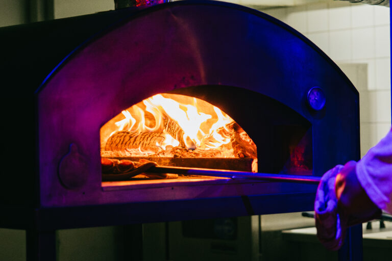 Een pizzaoven met vlammen en een hand die een metalen spatel erin steekt.