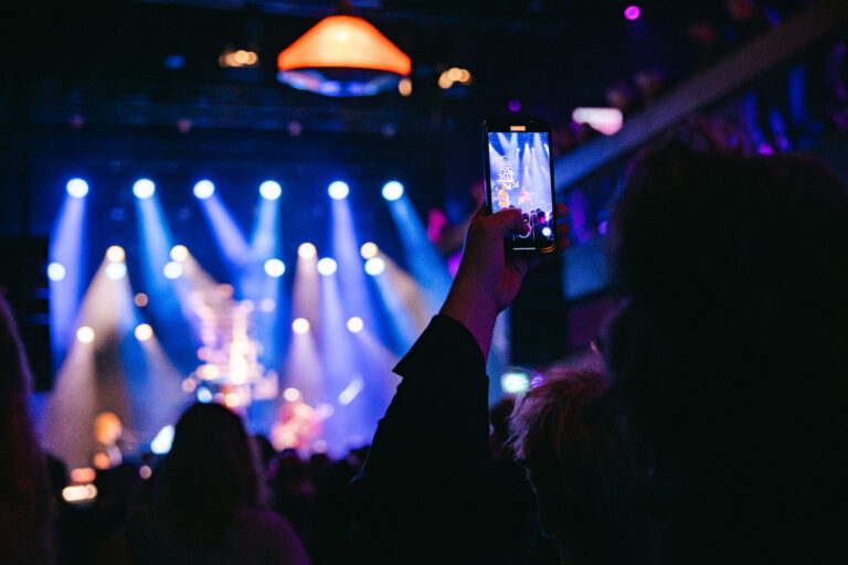 Een persoon houdt een smartphone omhoog om een optreden te filmen, met kleurrijke lichten op de achtergrond.