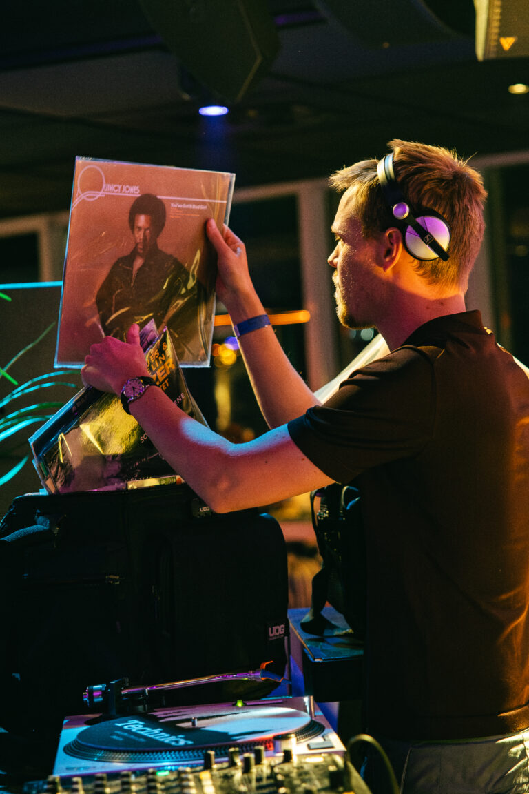 Een DJ staat achter een draaitafel en houdt een vinylplaat omhoog terwijl hij draait.