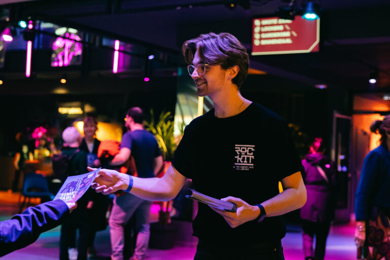 Een man in een zwart T-shirt deelt affiches uit op een evenement met kleurrijke verlichting en jonge mensen op de achtergrond.