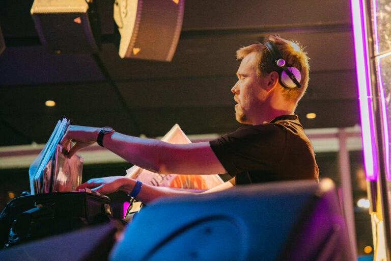 Een DJ selecteert vinylplaten terwijl hij aan het draaien is op een podium met kleurrijke verlichting.