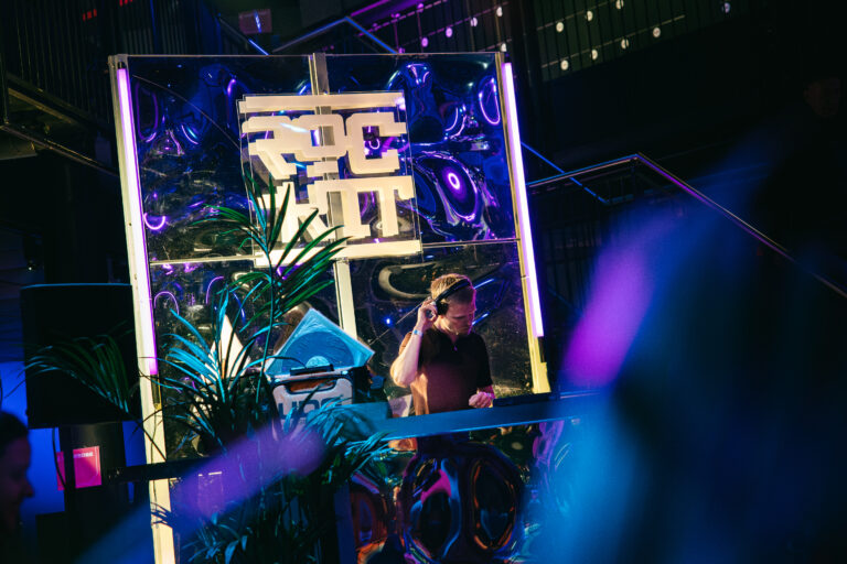 Een DJ staat achter een kleurrijke draaitafel met leuke verlichting in een levendige partyomgeving.